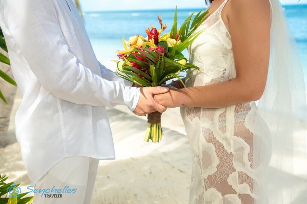 A pár egymás felé fordulva egymás kezét fogja, köztük a sárga és piros virágokból és pálmalevélből álló menyasszonyi csokor.