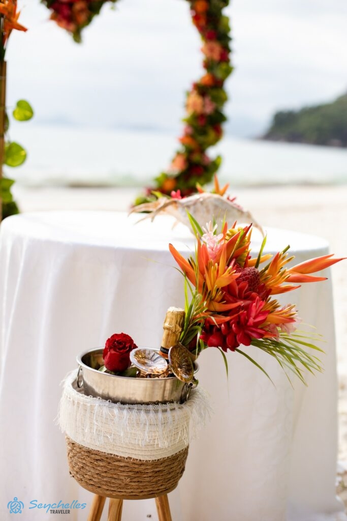 Trópusi virágokból készült, narancssárgás színekben pompázó menyasszonyi csokor a pezsgős vödörben Mahén.