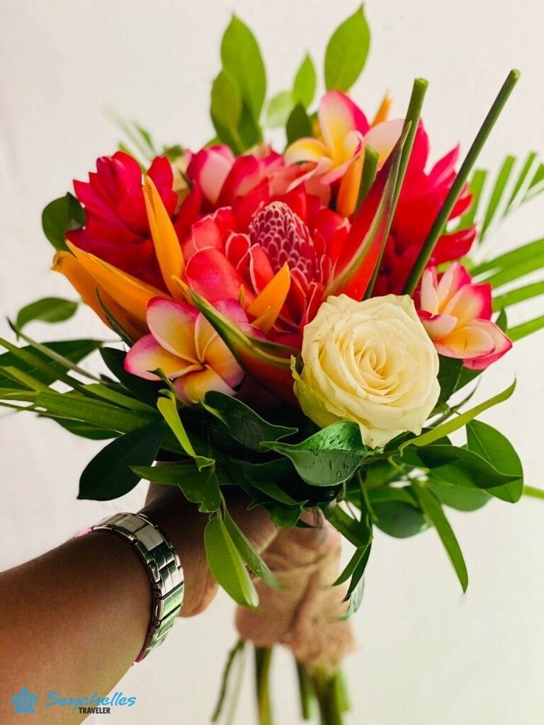 Pirosas-sárgás árnyalatú, trópusi virágokból és rózsából készült, zöld levelekkel díszített menyasszonyi csokor.