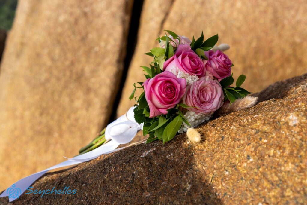Fehér szalaggal átkötött, rózsaszín rózsákból és fehér virágokból álló menyasszonyi csokor egy gránitsziklán Praslin-en.