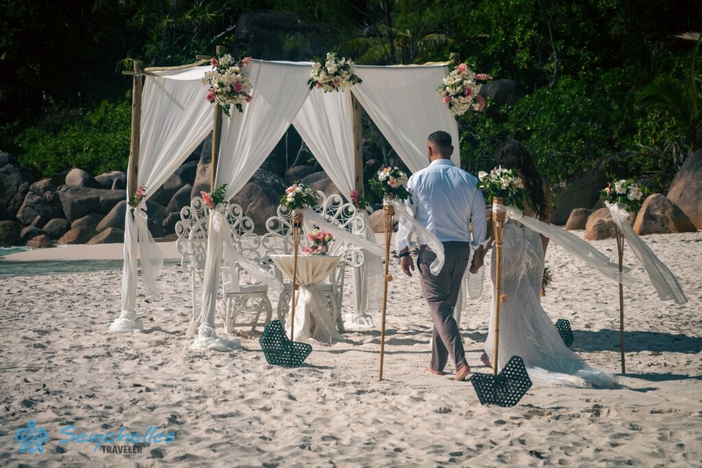 Az ifjú pár a 6 oszlopból álló bevonuló folyosón az esküvői pavilon felé lépdel Anse Georgette tengerparton.