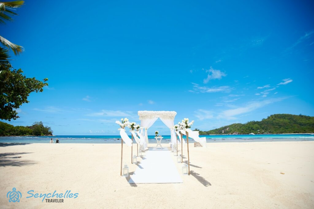 Fehér virágokkal sűrűn díszített esküvői pavilon és bevonuló folyosó bambusz oszlopokkal Baie Lazare tengerparton.