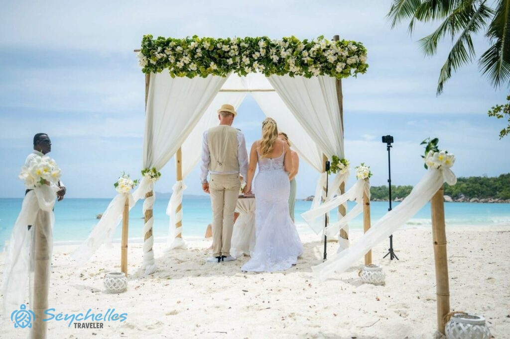 A jegyespár az anyakönyvvezetőt hallgatja a fehér virágokkal és zöld levelekkel díszített esküvői pavilon alatt Praslin-en.