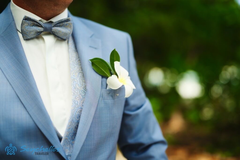 Fehér frangipániból és zöld levélből álló gomblyukdíszt visel a vőlegény Mahén.