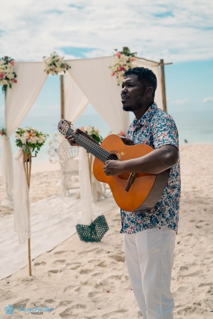 Egy gitáros férfi énekel Anse Georgette tengerparton a feldíszített esküvői helyszín mellett.