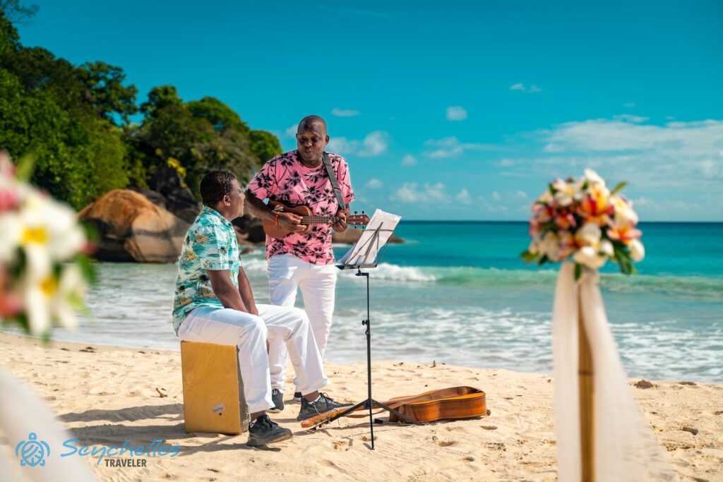 Két fehér nadrágos, színes inges zenész kottaállvánnyal és gitárokkal Anse Louis tengerparti esküvőn.