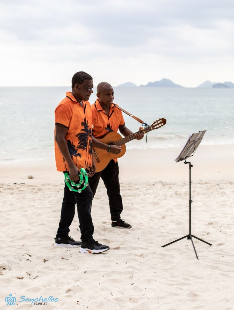 Két színes inges zenész kottaállvánnyal, az egyik gitárral, a másik kézi csörgő hangszerrel, Anse Louis tengerparton.