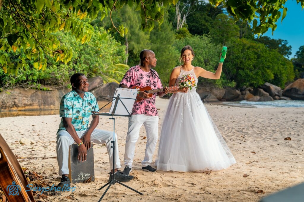 A menyasszony csörgőkarikát fog a kezében a két zenész mellett állva Anse Louis tengerparton.