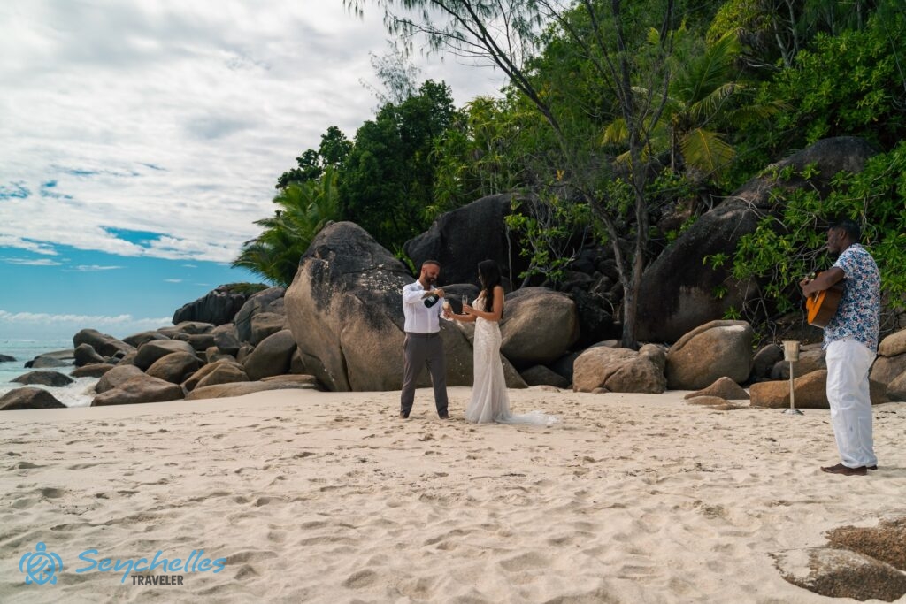 A férj habzóbort tölt feleségének a sziklák előtt állva Anse Georgette tengerparton, közelükben egy gitáros zenész.